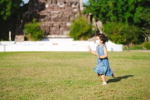 schattige Aziatische jongen meisje draait op gras in warme zon. kinderen dragen een gezichtsmasker om pm2.5 giftig stof te voorkomen en het virus te verspreiden. 5 jaar oud kind draagt blauwe spijkerjurk. foto