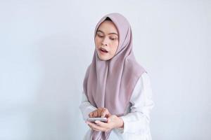 jonge aziatische islamvrouw die een hoofddoek draagt, is geschokt en wauw in de smartphone. Indonesische vrouw op grijze achtergrond foto