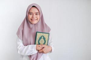 jonge aziatische islamvrouw die een hoofddoek draagt, houdt heilige al koran in de hand met een glimlach en een blij gezicht. Indonesische vrouw op grijze achtergrond foto