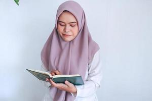 jonge aziatische islamvrouw die een hoofddoek draagt, bidt of leest de koran - het heilige boek van de islam met een glimlach en een serieus gezicht. Indonesische vrouw op grijze achtergrond foto