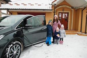 jonge vrouw met kinderen houdt eco-tassen vast en laadt elektrische auto op in de tuin van haar huis. foto