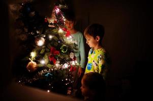 kinderen kijken op kerstboom met glanzende slingers op avond thuis. foto