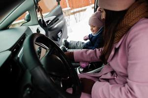 jonge moeder met dochtertje van de baby zit in de elektrische auto in de tuin van haar huis in de winter. foto