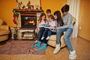 gelukkige jonge grote familie bij een open haard in warme woonkamer op winterdag. moeder met vier kinderen thuis lees boek. foto