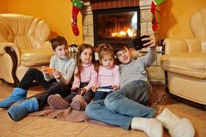 gelukkige vier kinderen met gadgets die thuis selfie-foto maken bij een open haard in een warme woonkamer op winterdag. foto