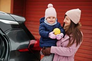 jonge moeder met babymeisje op handen tegen auto, houd citroen vast in de winter. foto
