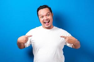 portret van dikke aziatische man in wit t-shirt glimlachend en wijzend naar zichzelf, vertrouwensconcept, close-up portret tegen blauwe achtergrond. foto