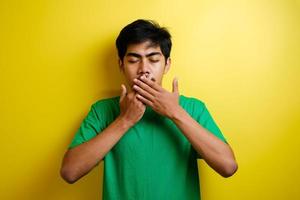 Aziatische jonge man met een groen t-shirt over een geïsoleerde gele achtergrond, sluit zijn ogen terwijl hij zijn mond bedekt met handen voor fouten foto