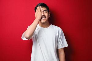 jonge aziatische man met een wit t-shirt glimlachend en lachend met de hand op het gezicht dat ogen bedekt voor verrassing. blind concept foto