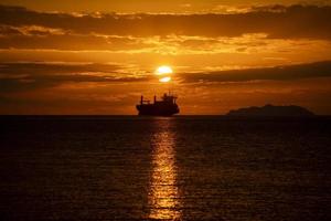 vrachtschip bij zonsondergang in tegenlicht met eiland op de achtergrond foto