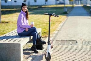 vrouwelijke student met elektrische scooter die een koffiepauze neemt, zittend op een bankje buiten haar college. foto