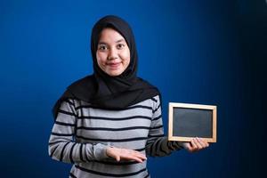 portret van slimme, succesvolle aziatische moslimvrouw die hijab draagt en naar de camera glimlacht terwijl ze een leeg schoolbord vasthoudt foto