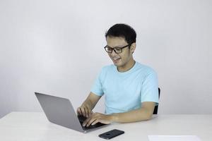 jonge aziatische man die zich gelukkig voelt en glimlacht wanneer een laptop op tafel werkt. Indonesische man met een blauw shirt. foto