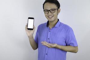 jonge aziatische man draagt blauw shirt staat en glimlachend wijzend op witte lege ruimte op smartphonescherm op witte achtergrond. foto