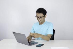 jonge aziatische man die zich gelukkig voelt en glimlacht wanneer een laptop op tafel werkt. Indonesische man met een blauw shirt. foto