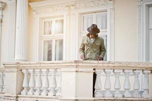 mode portret van zwarte Afro-Amerikaanse man op groene jas mantel en zwarte hoed, blijf op het balkon van geel herenhuis foto