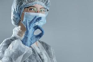 een lachend meisje in onscherpte in een medisch masker, rubberen handschoenen en wegwerpkleding vertoont teken ok. foto