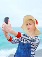 mooie jonge blonde blanke vrouw op vakantie in strohoed, gestreepte blouse en denim overall die selfie op smartphone maken door de verbazingwekkende blauwe zee-achtergrond foto