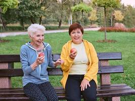 twee volwassen vrouwen die appels eten op de bank na het doen van sportoefeningen in het park. gezond levensstijlconcept foto