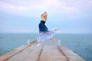 Kaukasische witte blonde zwangere vrouw met kort haar in lange jurk en sneakers blij om zwanger te zijn en bij de zee te springen in de zonsopgang foto