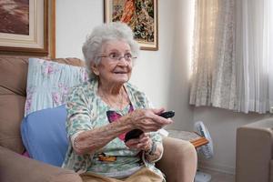 oudere dame thuis zittend in een stoel met een tv-zenderwisselaar in haar hand foto