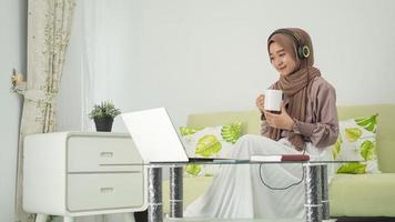 jonge vrouw in hijab die vanuit huis werkt en geniet van een drankje terwijl ze op een koptelefoon luistert foto