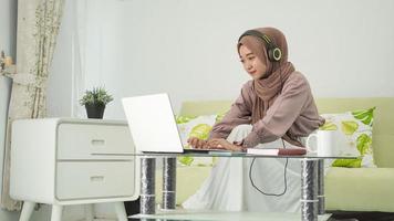 Aziatische vrouw in hijab die vanuit huis werkt en laptop typt terwijl ze luistert foto