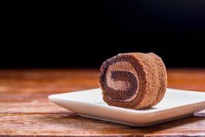 close-up heerlijke chocoladebroodjestaart met slagroom in een vierkante witte plaat op een houten tafel in de thuiskeuken met een zwarte achtergrond en kopieerruimte. zelfgemaakte bakkerij concept. foto