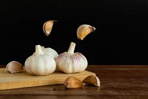 groep knoflook op snijplank en wat teentjes knoflook zwevend in de lucht op houten tafel met zwarte achtergrond. kopieer ruimte voor uw tekst. foto