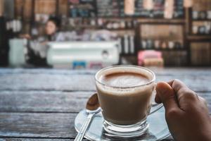 hand met een kopje koffie op houten tafel met wazig café achtergrond. koffietijden en ontspannend concept. kopieer ruimte voor uw tekst. foto