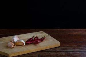 groep knoflook op snijplank en rode gedroogde chili op houten tafel met zwarte achtergrond. kopieer ruimte voor uw tekst. foto