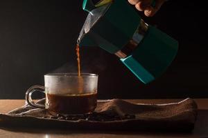 giet hete zwarte koffie uit de groene moka-pot om de koffiekop met rook en koffiebonen op bruin tafelkleed en houten tafel schoon te maken. voordeel van koffieconcept. foto