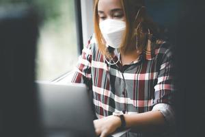 zakenvrouw die een masker draagt en op een laptop in een trein werkt, een aziatische freelance schrijver of ontwerper die e-mails schrijft op de computer, werkt en glimlacht op een comfortabele fauteuil in de buurt van het raam van de trein. foto