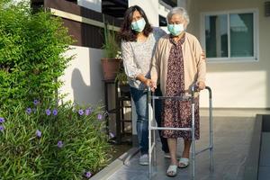 verzorger helpt aziatische senior of oudere oude dame vrouw lopen met rollator op helling en het dragen van een gezichtsmasker ter bescherming van de veiligheidsinfectie covid-19 coronavirus. foto