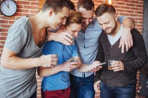 vrolijke oude vrienden communiceren met elkaar en telefoon kijken, glazen whisky in de pub. conceptentertainment levensstijl. wifi verbonden mensen in bartafelvergadering foto