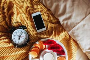 verschillende soorten fruit voor het ontbijt met klok die 7 uur aangeeft en smartphone op het bed foto