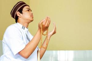 zijaanzicht van jonge aziatische moslim die tot allah bidt. islam man zit met bedelend handgebaar en houdt gebedskralen in zijn hand foto