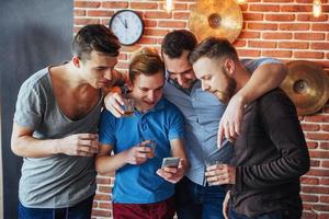 vrolijke oude vrienden communiceren met elkaar en telefoon kijken, glazen whisky in de pub. conceptentertainment levensstijl. wifi verbonden mensen in bartafelvergadering foto