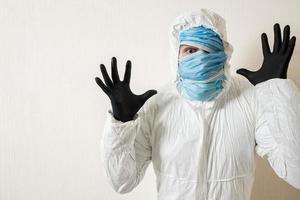 een bange man in een beschermend pak met medische maskers toont afschuw tegen een witte muur. de verschrikkingen van de epidemie, het gevaar van het coronavirus foto