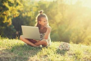 klein meisje praat op een laptop terwijl ze op het gras in de zon zit. gekleed in een sarafan en hoed foto