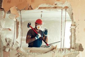 bouwer met een hamer in zijn handen breekt de betonnen muur de bouwer is gekleed in een beschermend pak en helm foto