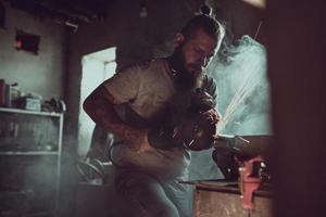 knappe brutale man met een baard die een motorfiets repareert in zijn garage die met een cirkelzaag werkt. in de garage veel vonken en rook van het zagen foto