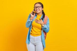 portret van vrolijke jonge Aziatische vrouw student in casual kleding met rugzak praten op smartphone met handen op borst geïsoleerd op gele achtergrond. onderwijs in universiteitsconcept foto