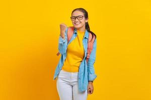 vrolijke jonge Aziatische vrouw student in denim outfit met rugzak staande doen winnende gebaar vieren geïsoleerd op gele achtergrond. onderwijs in universitair concept foto