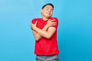 Portret van een vrolijke Aziatische jongeman in een rood shirt die zichzelf omhelst en het koud heeft geïsoleerd op een blauwe achtergrond foto