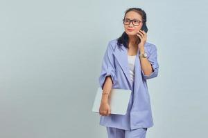 portret van glimlachende jonge aziatische vrouw die op mobiele telefoon spreekt en laptop houdt die op witte achtergrond wordt geïsoleerd foto