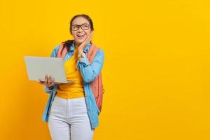 portret van opgewonden jonge Aziatische vrouw student in casual kleding met rugzak met behulp van laptop en wang aan te raken met handen geïsoleerd op gele achtergrond. onderwijs in universiteitsconcept foto