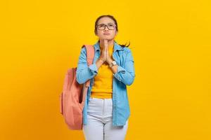 mooie jonge aziatische vrouw student in denim outfit met rugzak, handen gevouwen in gebed gebaar, bedelen over iets geïsoleerd op gele achtergrond. onderwijs in het concept van de hogeschool foto