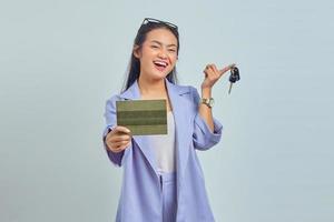 portret van vrolijke jonge aziatische vrouw die voertuigboek toont en voertuigsleutels houdt die op witte achtergrond worden geïsoleerd foto