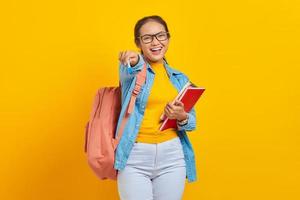 portret van vrolijke jonge Aziatische vrouw student in casual kleding met rugzak met boek en pen, kijkend naar camera geïsoleerd op gele achtergrond. onderwijs in universiteitsconcept foto